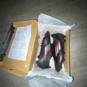 Adidas F50 svarta fotbollsskor - Säljer ett par svarta Adidas F50 fotbollsskor med röda och vita detaljer. Skorna har en slimmad design, platt sula med dobbar och är tillverkade i syntetmaterial för bästa grepp på planen. Perfekta för dig som vill sticka ut med stilrena och snabba skor.
