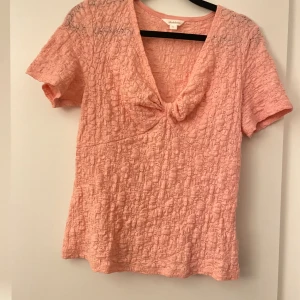 Rosa spetsblus med knut  - Säljer en ljusrosa blus i storlek S. Blusen har korta ärmar, v-ringad hals med snygg knutdetalj framtill och är gjord i ett spetsigt, broderat bomullstyg. Köpt på MQ! 