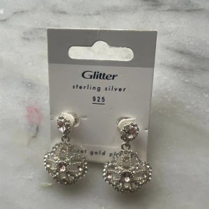 Glittriga örhängen i silver från Glitter - Snygga örhängen i äkta sterling silver 925 från Glitter. De har en rund, hängande design med glittrande stenar och ett detaljerat mönster.