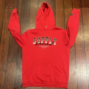 Röd hoodie Supply & Demand - Riktigt snygg röd hoodie från Supply & Demand med stor logga och text på bröstet. Tröjan har huva, känguruficka och en svartvit patch på ena ärmen. Perfekt för dig som gillar streetwear och vill sticka ut med en bold färg. Defekt: Hoodien saknar tyvärr snören.