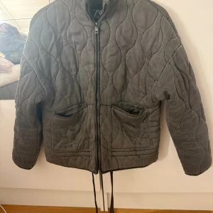 Quiltad grå jacka från Zara  - Quiltad grå jacka från Zara i storlek XS. Jackan har dragkedja framtill, två stora fickor och snörning i nederkant. Snygg vågig quiltning och hög krage ger en cool vibe. Perfekt för dig som gillar stilrena och trendiga plagg.