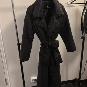 Svart lång puffer trenchcoat 157 - Svart lång puffer trenchcoat från 157 med bred krage och knytbälte i midjan. Jackan har en oversized look, långa ärmar och är tillverkad i glansig syntet. Perfekt för kalla dagar när du vill ha både stil och värme.