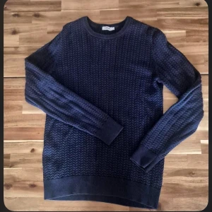 Mörkblå stickad tröja från J lindberg  - Säljer en mörkblå stickad tröja från J.lindberg med snyggt strukturmönster. Tröjan har rund halsringning och långa ärmar, perfekt för kyliga dagar. Passar dig som gillar klassisk och stilren look.
