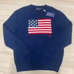 Mörkblå stickad tröja Polo Ralph Lauren - Mörkblå stickad tröja från Polo Ralph Lauren med amerikansk flagga broderad på bröstet och RL-detalj i rött. Tröjan har rund hals, ribbade muddar och långärmad passform. Perfekt för dig som gillar klassisk och stilren design.