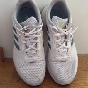 Adidas sportskor - Fräscha vita Adidas sneakers med mesh-ovandel, klassiska tre ränder i mörkgrönt och limegrönt på sidorna samt logga på plös och häl. Skorna har snörning och platt sula, perfekt för träning eller vardagsstil. Lätta och andas bra tack vare materialet.