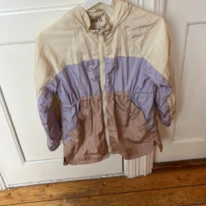 Pastellfärgad vindjacka från Zara - Snygg vindjacka från Zara i pastelliga färger med beige, lila och ljusbruna paneler. Jackan har huva, dragkedja framtill och elastiska muddar vid ärmsluten. Perfekt för blåsiga dagar och enkel att slänga på sig över en hoodie.