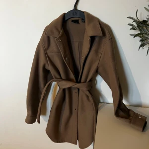 Brun kappa från Vero Moda  - Brun / mörkbeige kappa från Vero Moda med bälte i midjan och tryckknappar framtill. Jackan har klassisk krage, långa ärmar och är tillverkad i ett mjukt material som känns lite ulligt. Inga som helst defekter på jackan förutom att den kan uppfattas som lite nopprig.