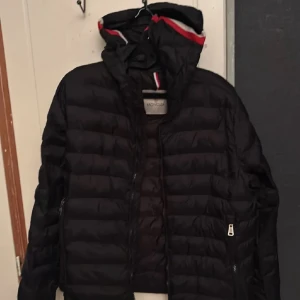 Svart pufferjacka från Holcola - Svart pufferjacka från Holcola med huva och dragkedja framtill. Jackan har horisontella quiltade partier och detaljer i rött och vitt på insidan av huvan. Perfekt för kyliga dagar och har praktiska fickor med dragkedja.