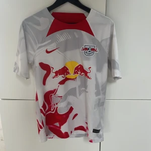 RB Leipzig vit och röd matchtröja Nike - Säljer en officiell RB Leipzig fotbollströja från Nike i vit med röda och grå detaljer, samt klubbens logga och Red Bull-tryck på bröstet. Tröjan har korta ärmar och är tillverkad i lätt Dri-Fit material som andas. RB Leipzig-tryck på ryggen och coolt grafiskt mönster.