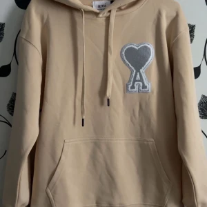 Beige hoodie från Ami Paris med hjärtdetalj - Snygg beige hoodie från Ami Paris med stor grå hjärtdetalj och bokstaven A på bröstet. Tröjan har huva med snörning, magficka och ribbade muddar. Tillverkad i mjuk bomull, perfekt för chill dagar. Stilren och trendig design som sticker ut.