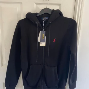 Svart hoodie från Polo Ralph Lauren - Svart hoodie från Polo Ralph Lauren med röd broderad logga på bröstet. Tröjan har huva med snörning, dragkedja framtill och två stora fickor. Tillverkad i mjuk bomull, perfekt för chill dagar. Klassisk och stilren design som funkar till jeans eller joggers.