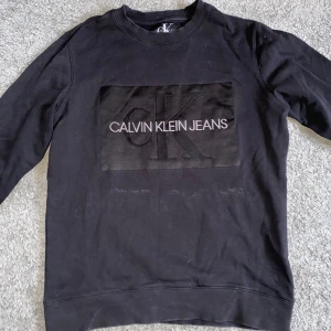 Svart sweatshirt från Calvin Klein Jeans - Svart sweatshirt från Calvin Klein Jeans med stort CK-logotyptryck på bröstet. Tröjan har rund hals, långa ärmar och ribbade muddar vid ärmslut och nederkant. Materialet är mjuk bomull som känns skön mot huden. Perfekt för en chill och stilren look.