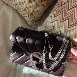 Sandro väska - Snygg axelväska i skinn med quiltad design och silverfärgad kedja som axelrem. Väskan har ett stort dekorativt metallspänne framtill och insidan är klädd i mjuk mocka. Perfekt storlek för det viktigaste och ger en cool vibe till din outfit.