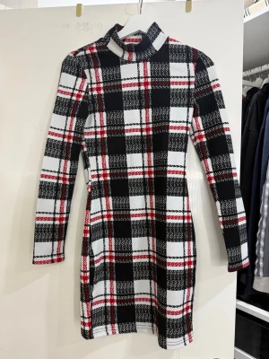 Rutig kortklänning med polokrage - Säljer en snygg kortklänning med svart, vit och röd rutig design. Klänningen har lång ärm och polokrage, vilket ger en cool och trendig vibe. Materialet känns mjukt och stretchigt, perfekt för dig som gillar att sticka ut med mönster.