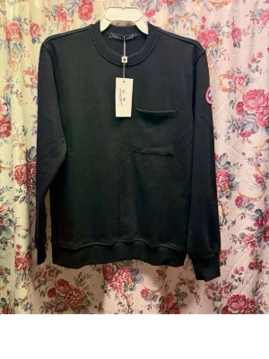 Svart sweatshirt från Canada Goose - Svart sweatshirt från Canada Goose med klassisk logga på ärmen och en bröstficka. Tröjan har rund halsringning och är tillverkad i en mjuk blandning av polyester och bomull. Perfekt för dig som gillar stilrena och bekväma plagg.