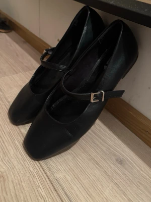 Svarta ballerinaskor med rem - Stilrena svarta ballerinaskor med fyrkantig tå och justerbar rem med silverfärgat spänne. Skorna har platt sula och är gjorda i ett syntetmaterial som liknar skinn. Perfekta för dig som gillar klassisk och enkel stil.