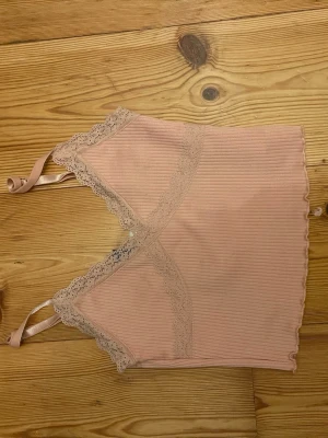 Rosa ribbad linnetopp från Divided XS - Supersöt ljusrosa ribbad linnetopp från Divided med spetsdetaljer längs v-ringningen och tunna axelband. Perfekt för sommaren och enkel att matcha med jeans eller kjol.