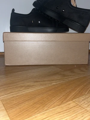 Svarta Christian Louboutin sneakers med nitar - Säljer ett par svarta sneakers från Christian Louboutin i mocka med svarta nitar på tån och den ikoniska röda sulan. Skorna har snörning och en clean, rund tå. Kommer med originalkartong och dustbag.