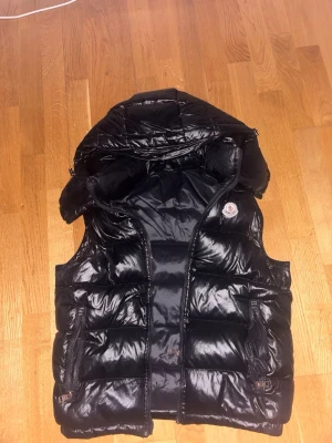 Svart dunväst från Moncler - Säljer en svart, glansig dunväst från Moncler med huva och dragkedja framtill. Västen har två fickor med dragkedja och ett klassiskt Moncler-märke på bröstet. Perfekt för lager-på-lager och har en quiltad design som ger extra värme. Obs litet hack i dragkedjan men syns knappast eller förstör nått 