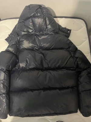 Svart Moncler pufferjacka med huva - Säljer en svart pufferjacka från Moncler med huva och dragkedja. Jackan har Moncler-logga på bröstet och coolt innerfoder med serietidningsmönster. Fylld med dun och har en glansig finish som ger en riktigt modern vibe. Perfekt för kalla dagar och riktigt snygg till streetwear.