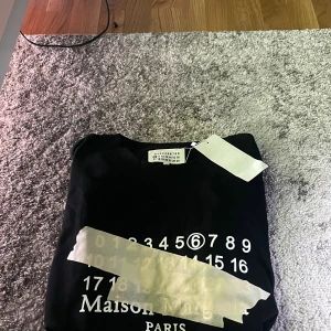 Maison Margiela svart t-shirt - Maison Margiela T-shirt med ikonisk tejpdetalj. Minimalistisk och stilren design med tydlig Margiela-estetik – perfekt för både vardag och lager-på-lager under höst/vinter.  ✔ Maison Margiela ✔ Bra skick (se bilder) ✔ Utan originalförpackning ✔ Klassisk passform  En eftertraktad t-shirt som funkar lika bra till jeans som under en jacka eller kavaj..