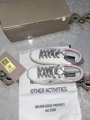 Golden Goose Super-Star sneakers vit/rosa - Golden Goose Super-Star sneakers i vitt skinn med grå snörning, grå stjärndetalj på sidan och ljusrosa hälparti. Klassisk rund tå och platt sula. Snygg kontrast mellan färgerna och ikonisk design som sticker ut.