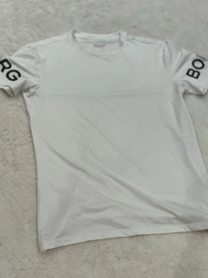 Vit t-shirt från Björn Borg - Säljer en stilren vit t-shirt från Björn Borg i storlek M. T-shirten har rund halsringning och svart text på ärmarna. Perfekt för en clean och sportig look. Materialet känns mjukt och lätt mot huden.