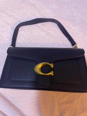 Svart axelväska från Coach i skinn - Säljer en svart axelväska från Coach i äkta skinn med en snygg guldig C-detalj på locket. Väskan har en stilren, rektangulär form och justerbart axelband. Perfekt för dig som gillar klassiska och trendiga accessoarer.