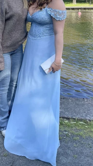 Ljusblå balklänning med spetsdetaljer - Säljer en ljusblå långklänning med vacker spets och broderade detaljer upptill. Klänningen har offshoulder-ärmar och en flowy kjol i tunt material. Perfekt för bal eller student. Snygg v-ringning och figurnära överdel som ger en elegant siluett. Jag köpte den ny för 5200 hos Carolinas bröllop, bal och gala i Vara.