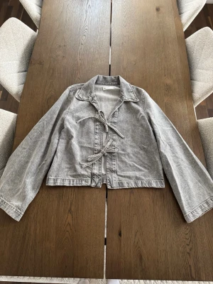 Grå jeansjacka med knyt från Gina Tricot - Säljer en grå jeansjacka från Gina Tricot i storlek S. Jackan/skjortan har två knytband framtill, klassisk krage och raka ärmar. Använd ett fåtal gånger, som ny. 