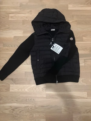 Svart Moncler cardigan - Säljer en svart pufferjacka från Moncler i storlek M. Jackan har stickade ärmar, quiltad kropp och en stor huva. Klassisk Moncler-logga på ärmen och dragkedja framtill. Perfekt för dig som vill ha en stilren och varm jacka med exklusiv känsla.