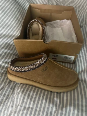 Oanvända UGGs  - UGGs i beige mocka med fluffigt foder och brunt mönster runt öppningen. Sulan är rejäl och greppvänlig, perfekt för kalla dagar. Klassisk UGG-logga på sidan och insidan är fodrad med mjuk päls.    Storlek 38! Box medföljer!