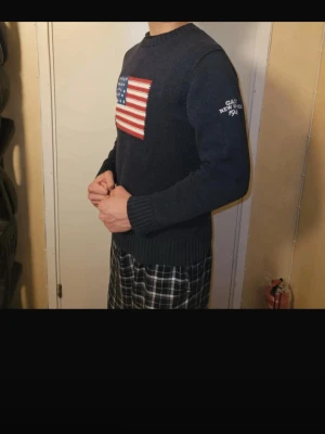 Mörkblå stickad tröja från Gant - Mörkblå stickad tröja från Gant med amerikansk flagga på bröstet och texten 'GANT NEW HAVEN 1949' på ärmen. Tröjan har rund hals, ribbade muddar och är långärmad. Perfekt för dig som gillar klassisk college-stil.