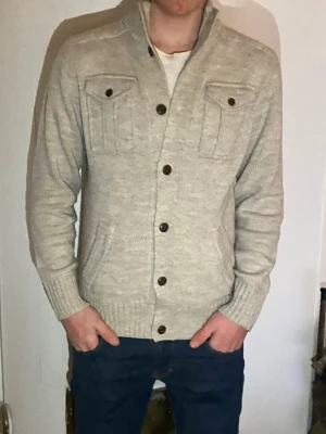 Ljusgrå kofta från Blue Inc - Säljer en ljusgrå stickad kofta från Blue Inc med ribbad krage och bröstfickor med knappar. Koftan har stora knappar framtill, två sidofickor och långärmad design. Perfekt för kyliga dagar och enkel att matcha med jeans.