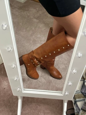 Bruna boots med nitar Micheal Kors - Snygga höga boots i brunt skinn med dekorativa guldfärgade nitar längs sidan och ett coolt spänne vid ankeln. Bootsens klack är stabil och ger lite extra höjd. Perfekta till höstlooken och matchas lätt med både jeans och kjol. Ifrån Micheal Kors!