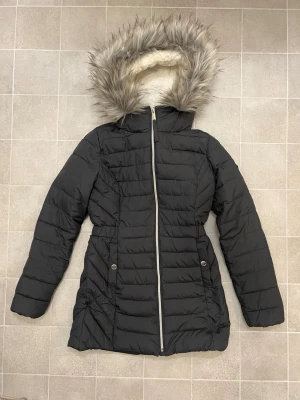 Svart pufferjacka med fuskpäls från Hollister - Mörkgrå lång jacka från Hollister med fluffig fuskpäls på huvan och mysigt vitt teddyfoder. Storlek XS. Jackan har dragkedja framtill, två fickor med knapp och snyggt Hollister-märke på ärmen. Perfekt för kalla dagar. Fuskpälsen är avtagbar. Har några lösa trådar men är i övrigt fint skick.