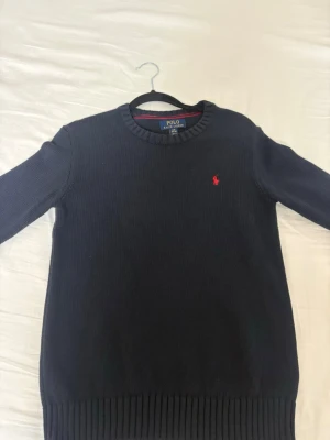 Mörkblå stickad tröja Polo Ralph Lauren - Klassisk mörkblå stickad tröja från Polo Ralph Lauren med rund halsringning och den ikoniska röda logon broderad på bröstet. Tröjan har ribbade muddar vid ärmslut och nederkant, perfekt för lager på lager under kyliga dagar. Tröjan passar ej mig och har inte lämnat tillbaka den, priset är ej hugget i sten så hör av dig!