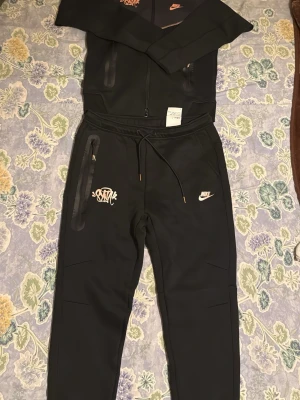 Nike mjukisset / tracksuit – Hoodie + byxor - Säljer ett riktigt snyggt och modernt Nike-set i svart med stilrena detaljer. Setet består av hoodie med dragkedja och matchande byxor. Båda plaggen har Nike-loggor samt en unik graffiti-inspirerad Nike-design som ger en exklusiv look.