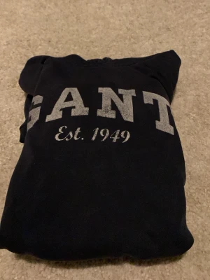 Gant hoodie  - Snygg och bekväm hoodie från GANT i klassisk mörk färg. Tröjan har trycket “GANT Est. 1949” på bröstet och en tidlös design som passar till det mesta. Mjuk och skön kvalitet, perfekt för både vardag och fritid.