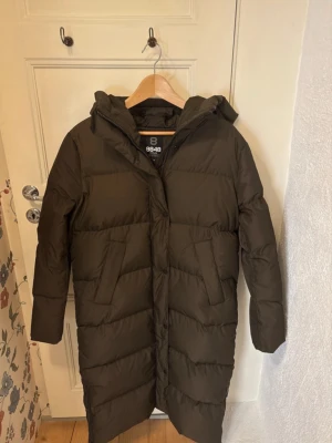 Brun lång dunjacka från 8848 Altitude - Säljer en lång, brun dunkappa från 8848 Altitude med huva och stora fickor. Jackan är quiltad och har en stilren look med diskret logga på ärmen. Perfekt för kalla dagar när du vill hålla dig varm och snygg. Modellen heter Biella 2.0 W Coat. Nypris 4200 kr. Säljer då den inte kommer till användning längre. Ett litet hål på jackans baksida vid midjehöjd, lätt att fixa men räknar med detta i priset!