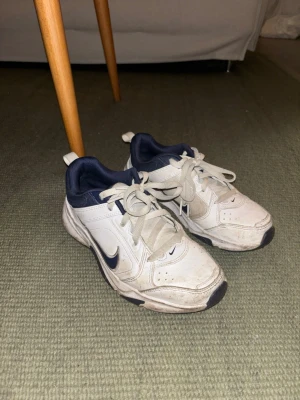 Vita och marinblå Nike sneakers - Nike sneakers i vitt och marinblått med klassisk swoosh på sidan. Skorna har snörning, rund tå och platt sula. Ovandelen är i skinn och syntet med meshdetaljer för extra komfort. Perfekta för dig som gillar sportig stil. Strl 39