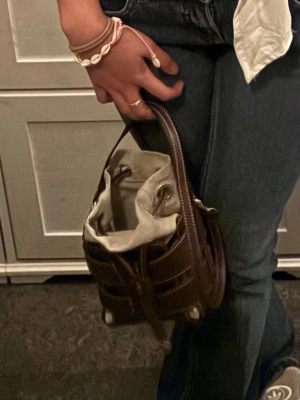 Brun handväska i skinn med snörning - Säljer en brun handväska i skinn med snygga detaljer och snörning upptill. Väskan har ett unikt flätat mönster på utsidan och handtag som gör den enkel att bära. Insidan är ljus och har en tygpåse som stängs med snöre. Perfekt för dig som vill ha en stilren och trendig accessoar.