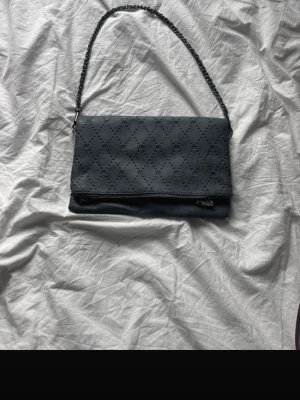 Svart axelväska med kedjerem - Säljer en svart axelväska från Fiorelli med snyggt perforerat mönster och kedjedetaljer på remmen. Väskan har dragkedja både fram och bak samt vikbar design. Perfekt storlek för det viktigaste och ger en cool vibe till din outfit.