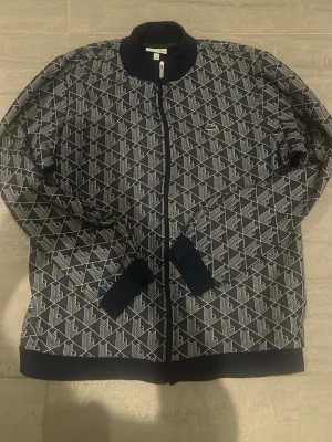 Mönstrad bomberjacka från Lacoste - Säljer en snygg mörkblå bomberjacka från Lacoste med vitt geometriskt mönster över hela jackan. Klassisk croc-logo på bröstet, ribbade muddar och dragkedja framtill. Perfekt för dig som vill sticka ut med stilren design.