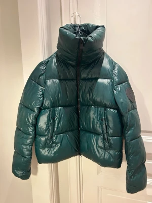 Grön pufferjacka från Save The Duck - Säljer en mörkgrön pufferjacka från Save The Duck med hög krage och två fickor framtill. Jackan är  helt animal free. Perfekt för kalla dagar och är jättevarm. Sparsamt använd och säljs då jag flyttar till ett varmt land. Jag har storlek S/M i normalt fall och jackan sitter perfekt.