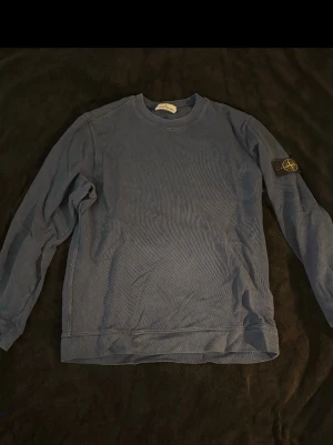 Blå Stone Island sweatshirt - Säljer en blå sweatshirt från Stone Island med klassisk rund hals och lång ärm. Tröjan har den ikoniska Stone Island-patchen på vänster ärm och är tillverkad i mjuk bomull. Perfekt för dig som gillar streetwear och vill ha en clean look. 