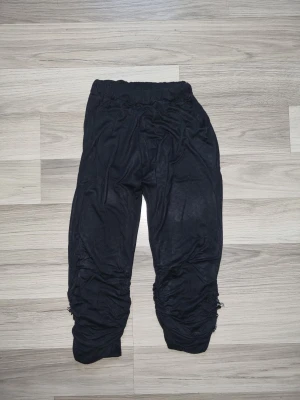 Svarta 3/4 leggings med strassdetaljer - Säljer ett par svarta 3/4 leggings med snygga strassdetaljer längst ner vid benen. De har en tight passform, elastisk midja och är gjorda i mjukt material. Benen är rynkade och dekorerade med glittrande stenar som ger en unik touch.