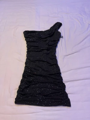 Svart glittrig one shoulder klänning - Säljer en svart kortklänning med glittrigt mönster och oneshoulder-design. Klänningen har en tight passform och är gjord i ett stretchigt syntetmaterial. Perfekt för dig som vill sticka ut med lite extra glitter och cool asymmetrisk axel.