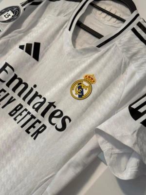 Real Madrid matchtröja Mbappé 9 - Säljer en vit Real Madrid matchtröja från Adidas med Mbappé och nummer 9 på ryggen. Tröjan har svarta detaljer, klubbmärke på bröstet, Champions League-märke på ärmen och 'Emirates Fly Better' tryckt framtill. Tillverkad i lätt polyester med diskret mönster.