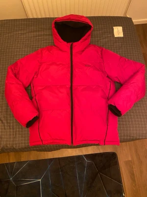Röd pufferjacka från BOSS - Snygg röd pufferjacka från BOSS med stor logga på ryggen och mindre logga på bröstet. Jackan har huva, svarta detaljer och hel dragkedja framtill. Insidan är svart med BOSS-mönster och jackan är fodrad för extra värme. Perfekt för kalla dagar. Köpt från Zalando och nypris ligger på 2200kr. Bara att skriva vid frågor och priset går såklart att diskutera!